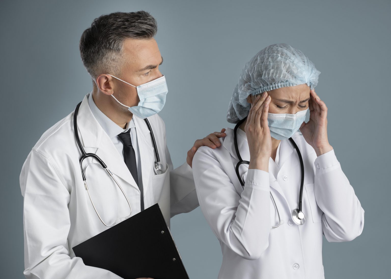 A imagem apresenta um médico mais velho de jaleco e camisa branca com uma gravata preta e uma médica de toca e jaleca com as mão na cabeça, os dois estão utilizando o estetoscópio e máscara