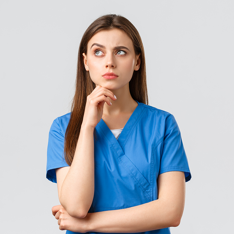 Médica confusa com a mão no queixo vestindo uniforme hospitalar azul em frente ao fundo cinza.