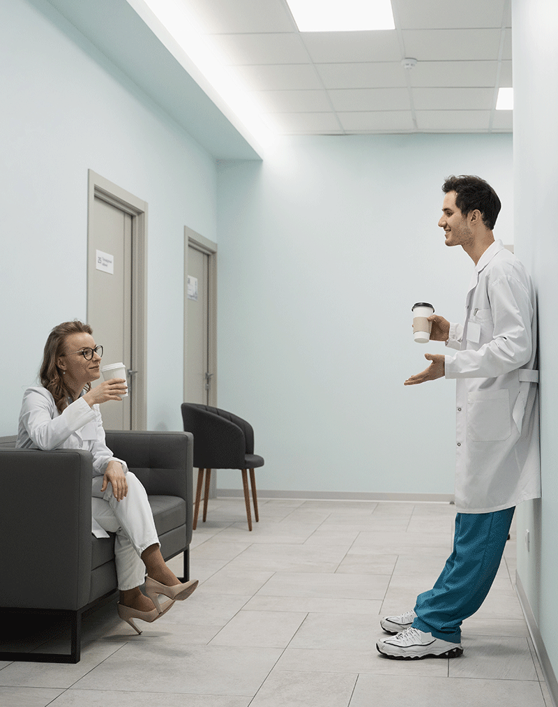 A imagem apresenta uma médica sentada na poltrona de uma hospital com uma xícara na mão, conversando um médico em pé a sua frente encostado na parede tomando café, ambos estão em corredor de hospital de cores neutras, vestindo jaleco e uniforme azul