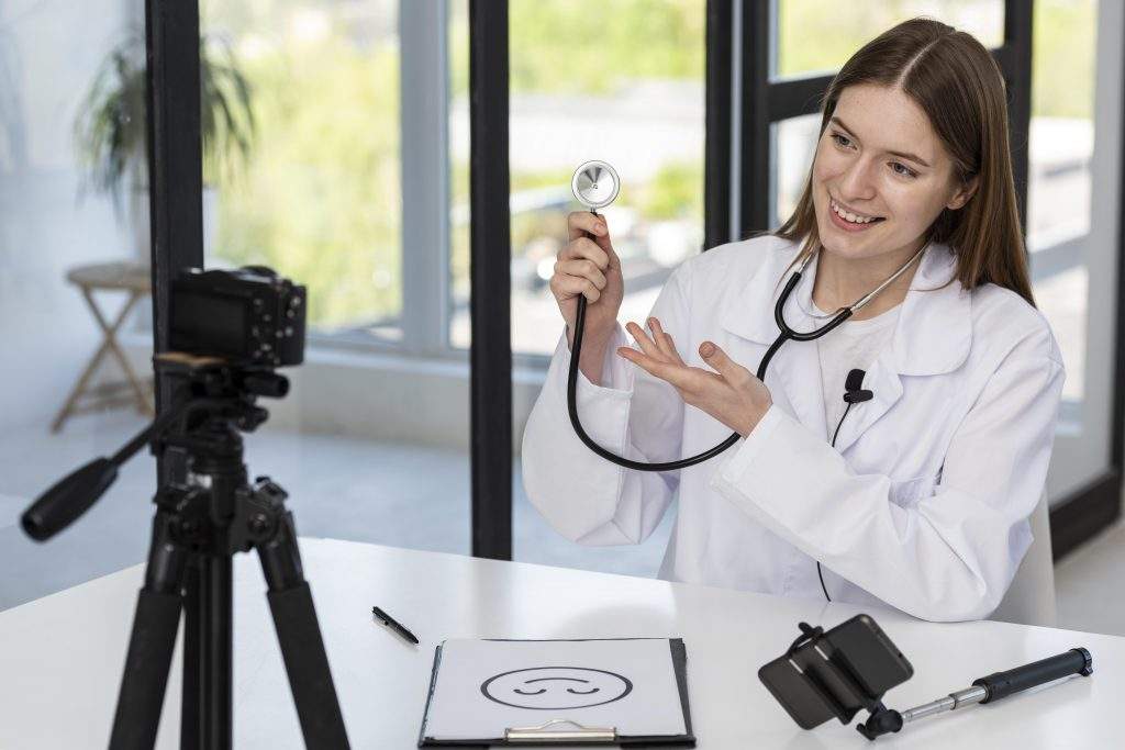 A imagem apresenta uma médica segurando um estetoscópio apontando para a câmera, ela está gravando conteúdo sentando em uma mesa em um ambiente claro com janelas amplas.