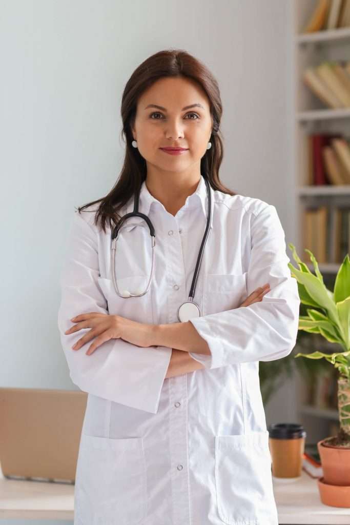 Médica empreendedora de braços cruzados, vestindo jaleco branco e com estetoscópio no pescoço em frente ao seu consultório médico